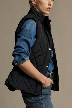 A person in a blue denim shirt and blue jeans wears the RM Williams Ikara Vest, a black quilted vest with snap buttons. Their right hand is in their jeans pocket as they stand sideways, with their face partially visible.