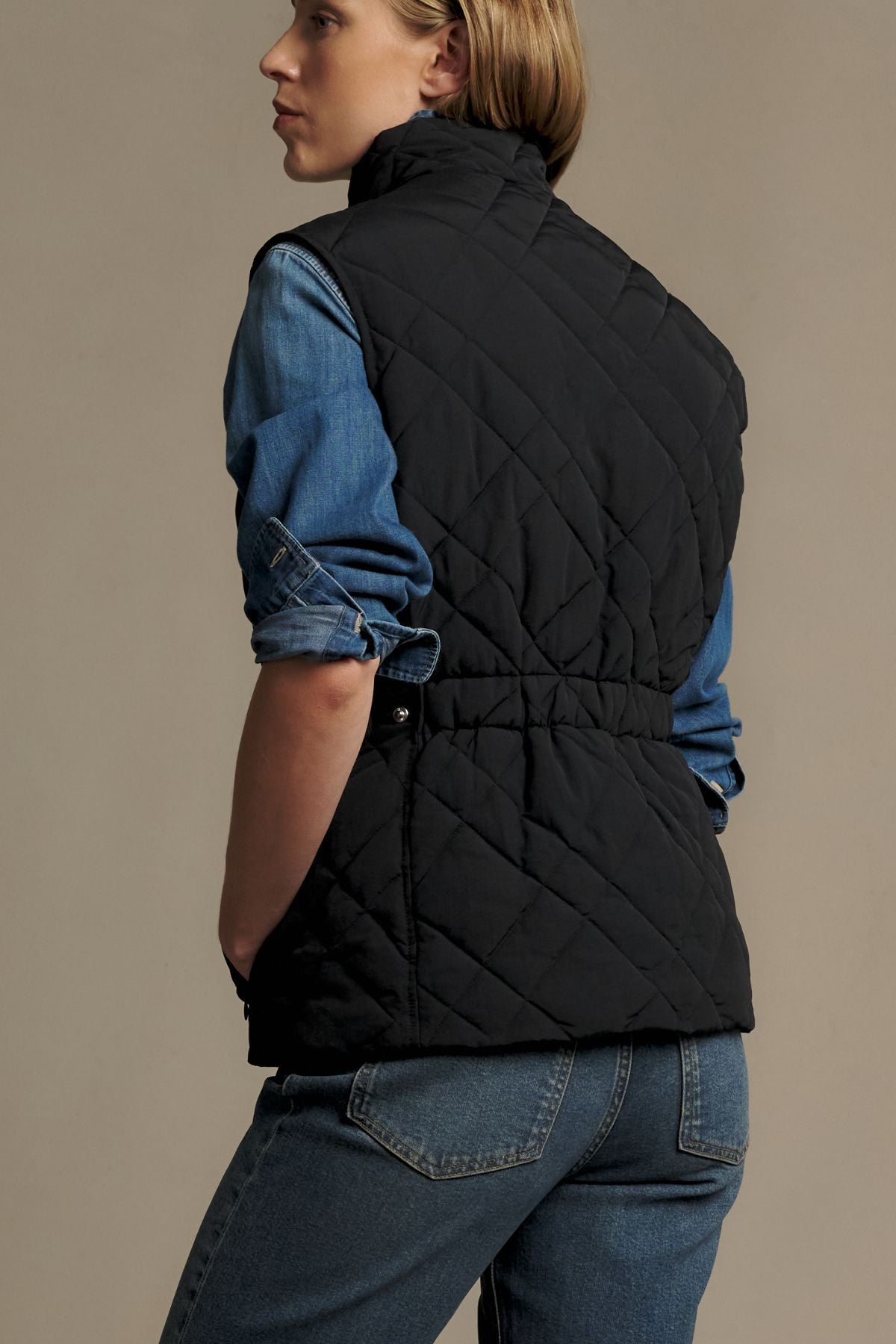 Wearing the RM Williams Ikara Vest in black over a blue denim shirt and blue jeans, a person stands facing away with their left hand in their pocket against a plain background.