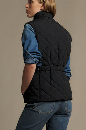 Wearing the RM Williams Ikara Vest in black over a blue denim shirt and blue jeans, a person stands facing away with their left hand in their pocket against a plain background.