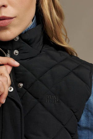 A woman wearing the RM Williams Ikara Vest—a black quilted vest with snap buttons—over a denim shirt holds the collar. Only the lower half of her face and torso are visible.