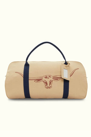 The RMW Nanga Canvas Bag features a tan, duffel-style design with a dark blue handle and strap. A prominent brown longhorn logo adorns the side, alongside a small tag on one handle highlighting its multi-purpose functionality.