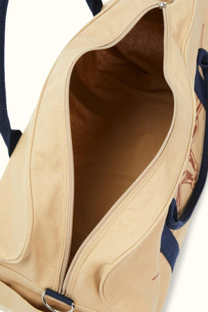 The RMW Nanga Canvas Bag boasts a beige duffle-style design with navy straps and a partially open zipper, showcasing its spacious interior. A metal ring on one corner adds versatility to its classic look.