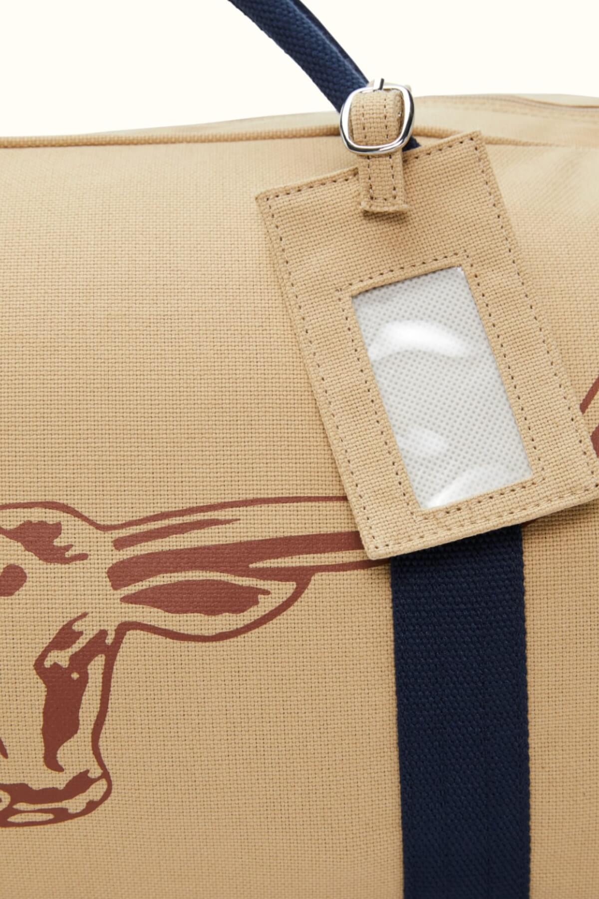 Close-up of the RMW Nanga Canvas Bag in tan, showcasing a partial brown longhorn steer image. This versatile duffel includes a black strap and a tan luggage tag with a clear window attached.