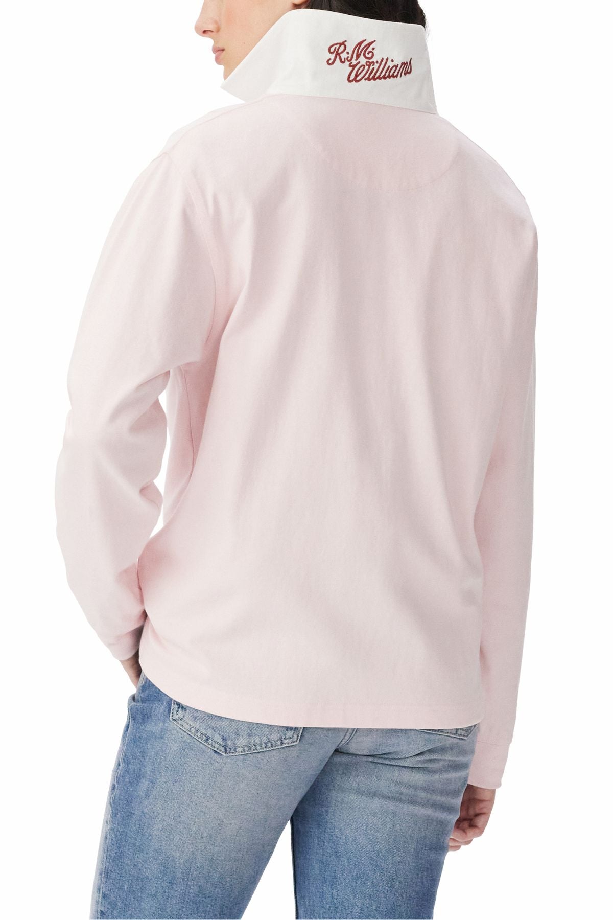 A person is shown from the back, hands in jean pockets, wearing the RM Williams Narran Heritage Rugby—a light pink organic cotton shirt with brushed twill collar and red "R.M. Williams" embroidery—paired with blue jeans.