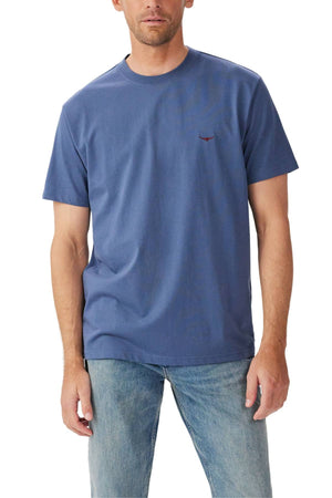 A man stands facing forward with his arms relaxed by his sides, wearing the RM Williams Parson T-Shirt in blue and light blue jeans against a plain white background.