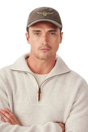 A man stands with arms crossed, wearing a beige half-zip sweater and the RM Williams Longhorn Twill Cap in gray with an adjustable strap, looking directly at the camera against a white background.