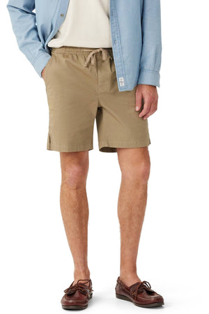 A person wearing RM Williams Washed Rugby Shorts with an elastic waistband and front slanted pockets, a white shirt, a light blue button-up shirt, and brown boat shoes stands with one hand in their pocket against a white background.