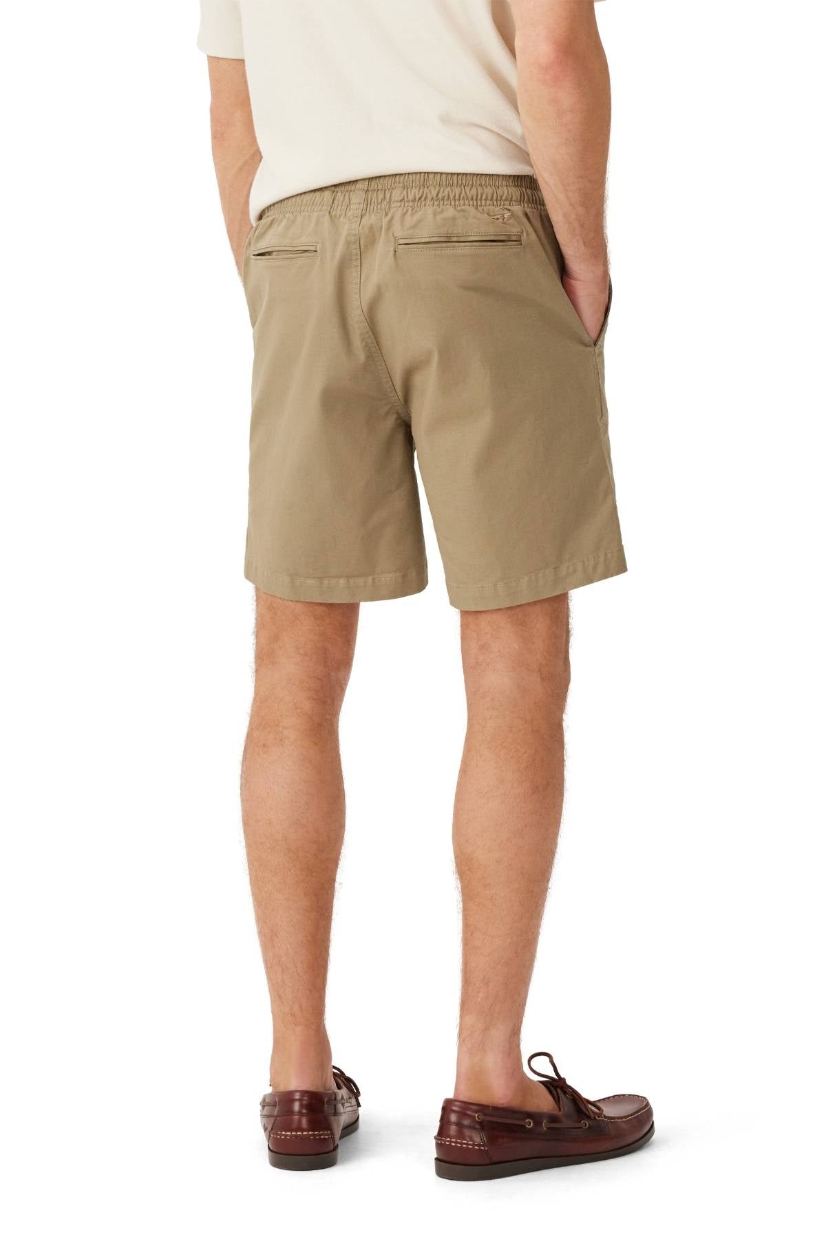 A man stands facing away, hands in his pockets, wearing a cream short-sleeve shirt, brown loafers, and RM Williams Washed Rugby Shorts with an elastic waistband, front slanted pockets, and branded embroidery. His legs are slightly apart.