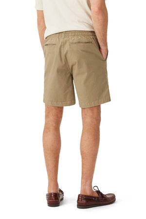 A man stands facing away, hands in his pockets, wearing a cream short-sleeve shirt, brown loafers, and RM Williams Washed Rugby Shorts with an elastic waistband, front slanted pockets, and branded embroidery. His legs are slightly apart.