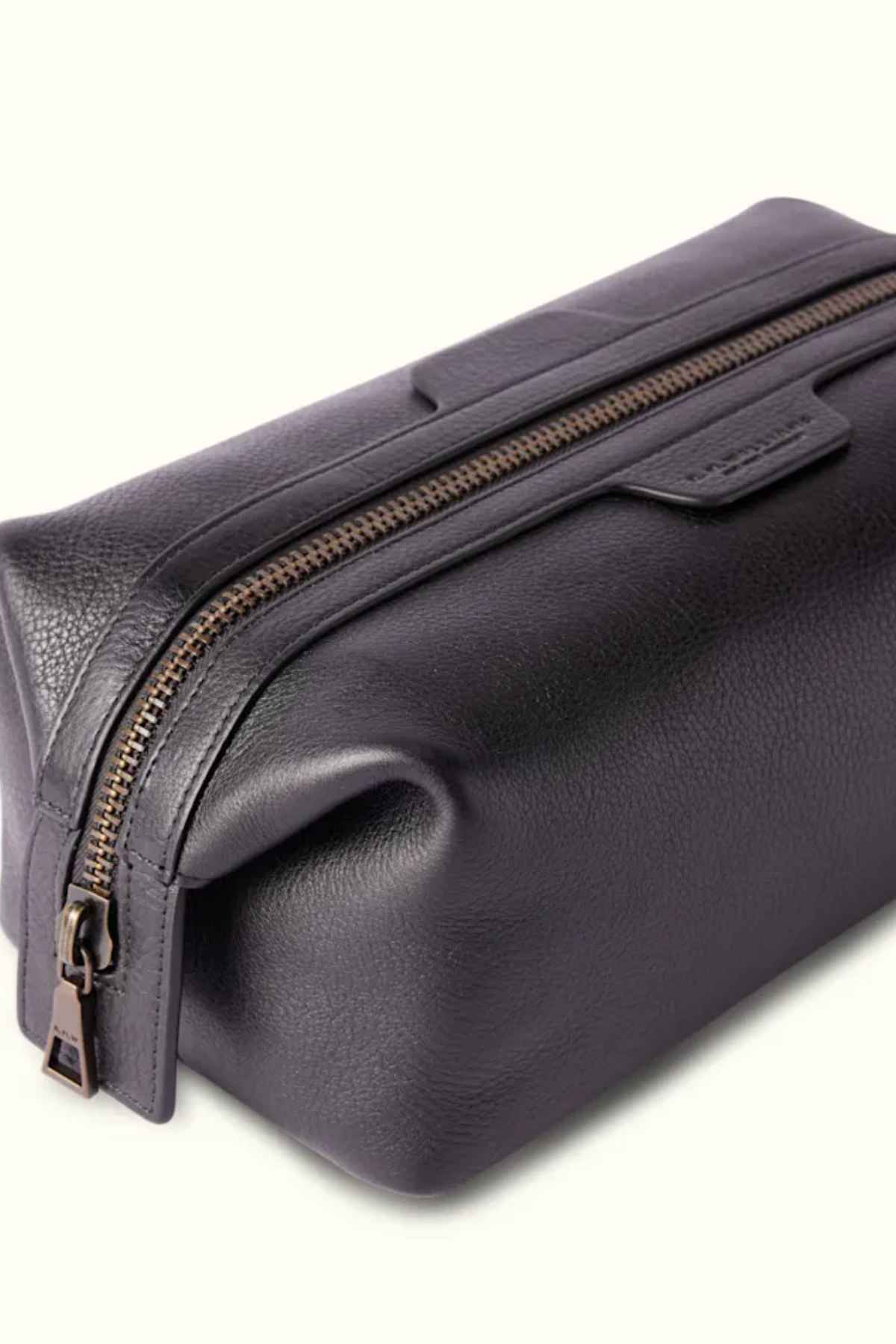 The RM Williams Farrier Washbag is crafted from dark leather with a smooth texture, featuring a frog-mouth zipper along the top. It has a rectangular shape and includes a small pull tab for easy opening and closing.