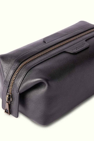 The RM Williams Farrier Washbag is crafted from dark leather with a smooth texture, featuring a frog-mouth zipper along the top. It has a rectangular shape and includes a small pull tab for easy opening and closing.