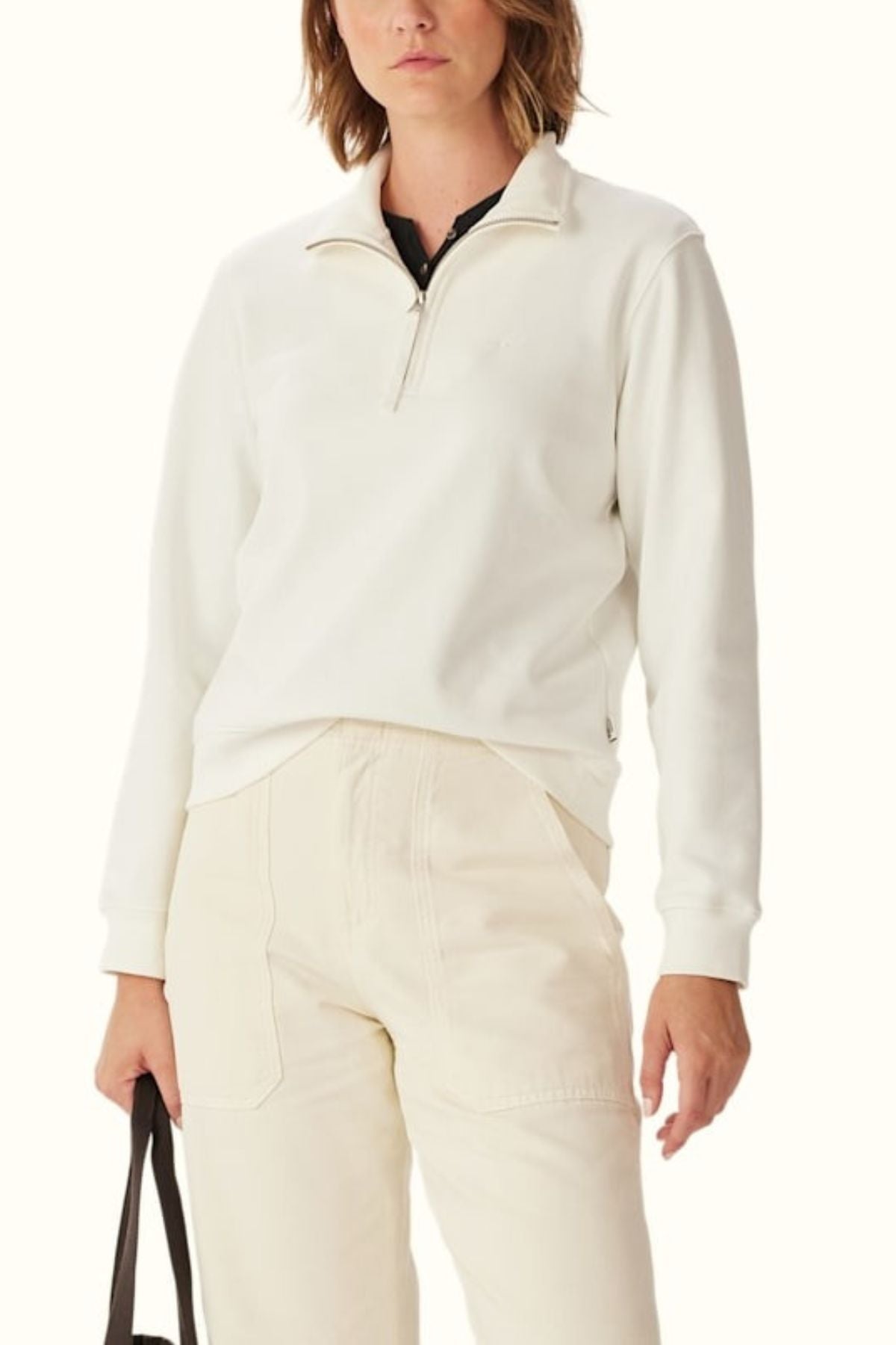 Wearing the RM Williams Trickett Zip Neck Sweatshirt in off-white with high-waisted cream pants and holding a black bag, cropped from shoulders down against a plain light background.