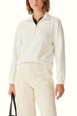Wearing the RM Williams Trickett Zip Neck Sweatshirt in off-white with high-waisted cream pants and holding a black bag, cropped from shoulders down against a plain light background.