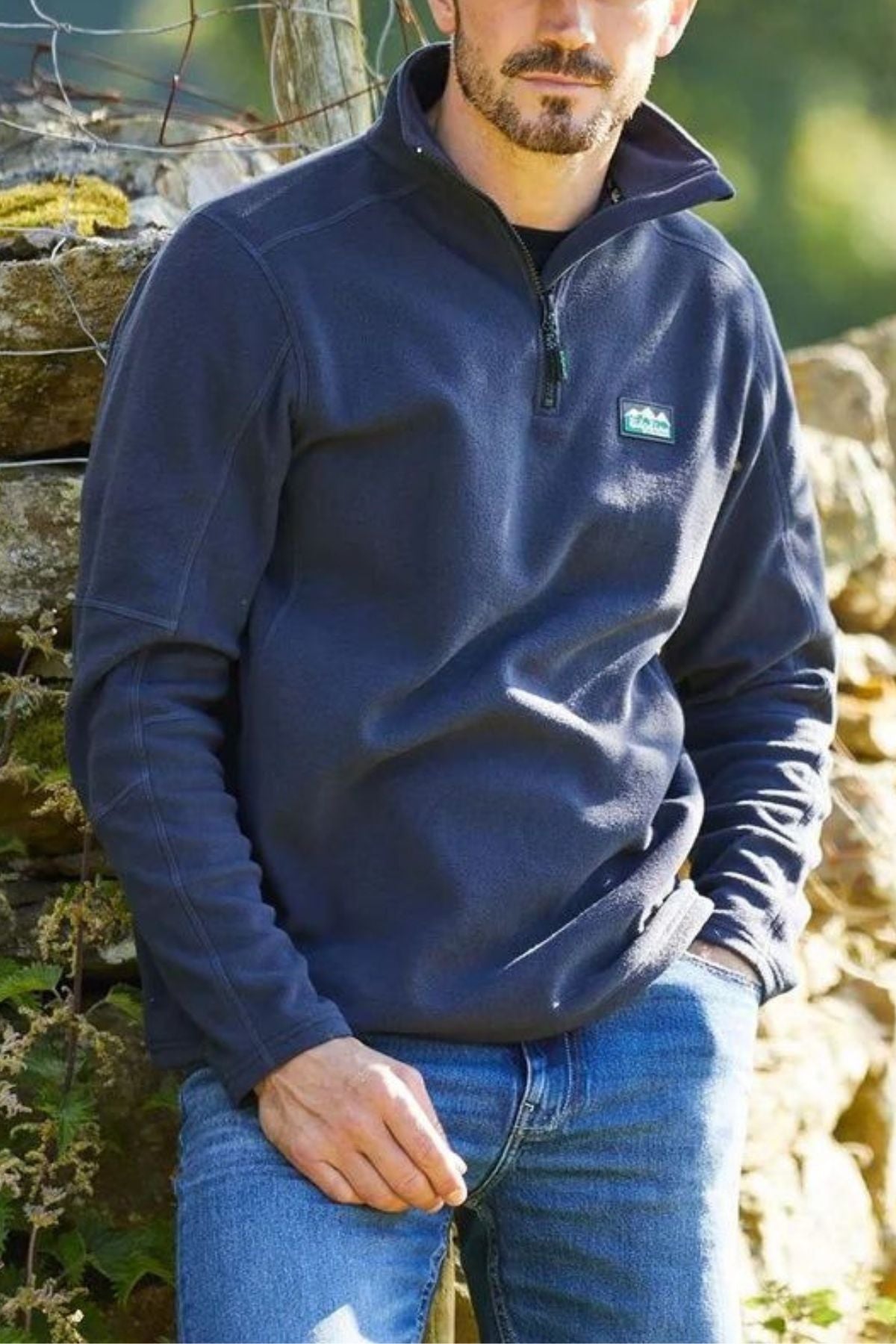 A man with a short beard wears a navy blue Ridgeline Narvik Fleece zip-up and blue jeans, sitting outdoors by a stone wall with greenery—ideal for outdoor performance.