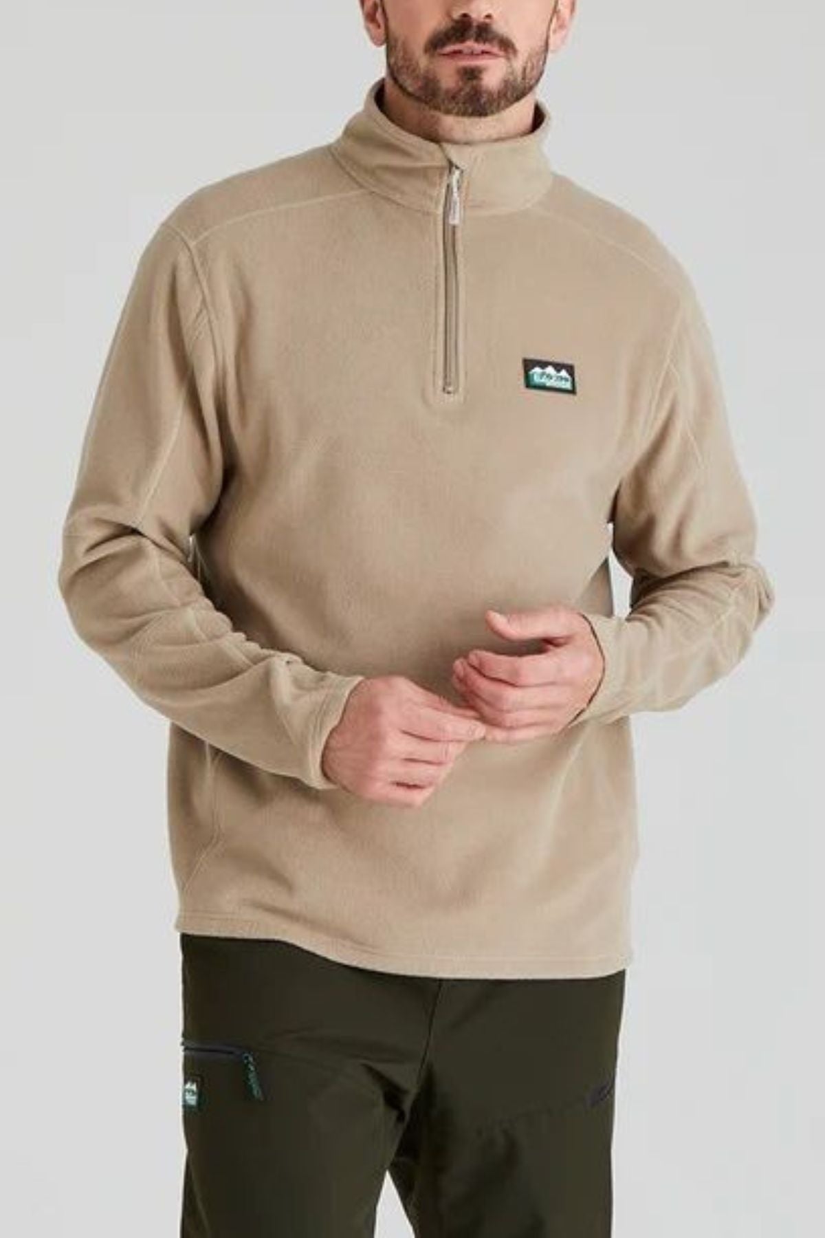A man wearing a Ridgeline Narvik Fleece lightweight zip-neck pullover with a small chest logo and dark green pants stands against a plain light background.