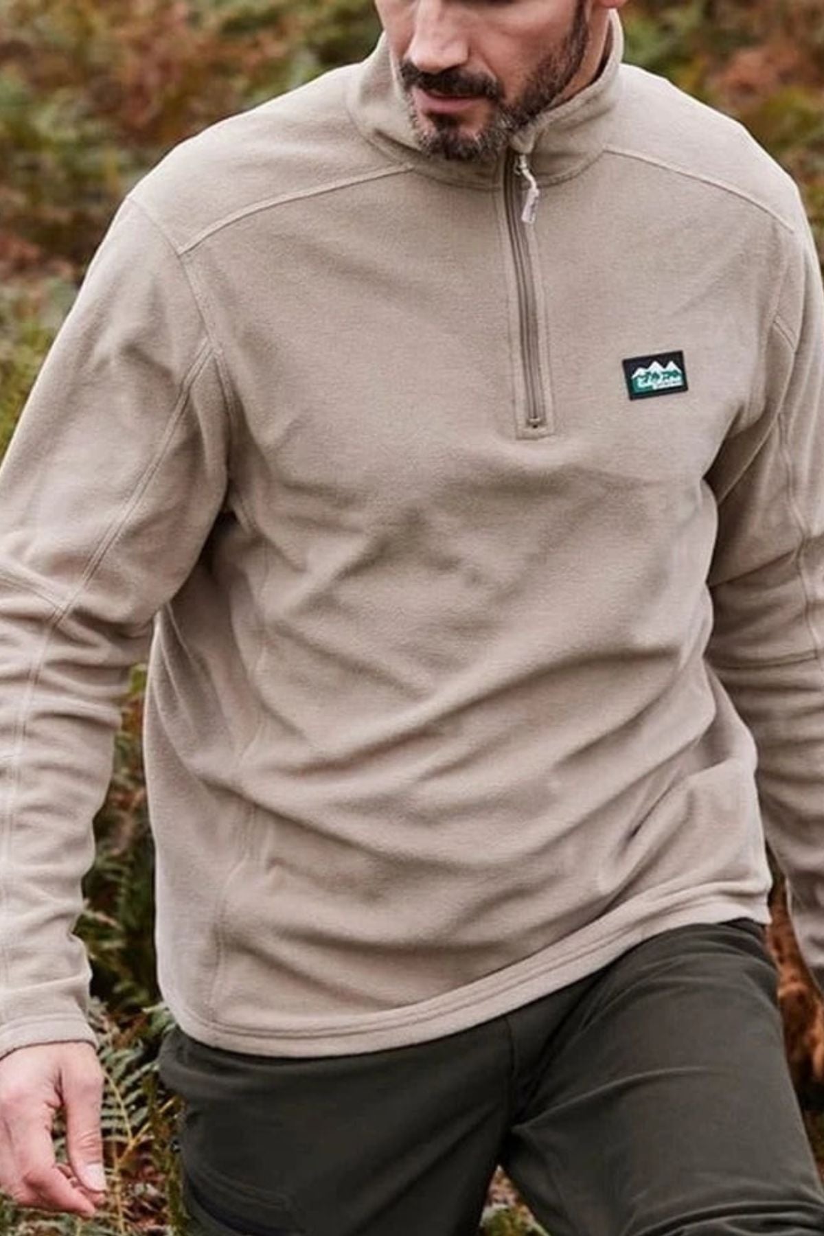A man wearing a Ridgeline Narvik Fleece with a zip-neck and small chest logo is walking outdoors through a wooded area, with only his upper body and part of his face visible.