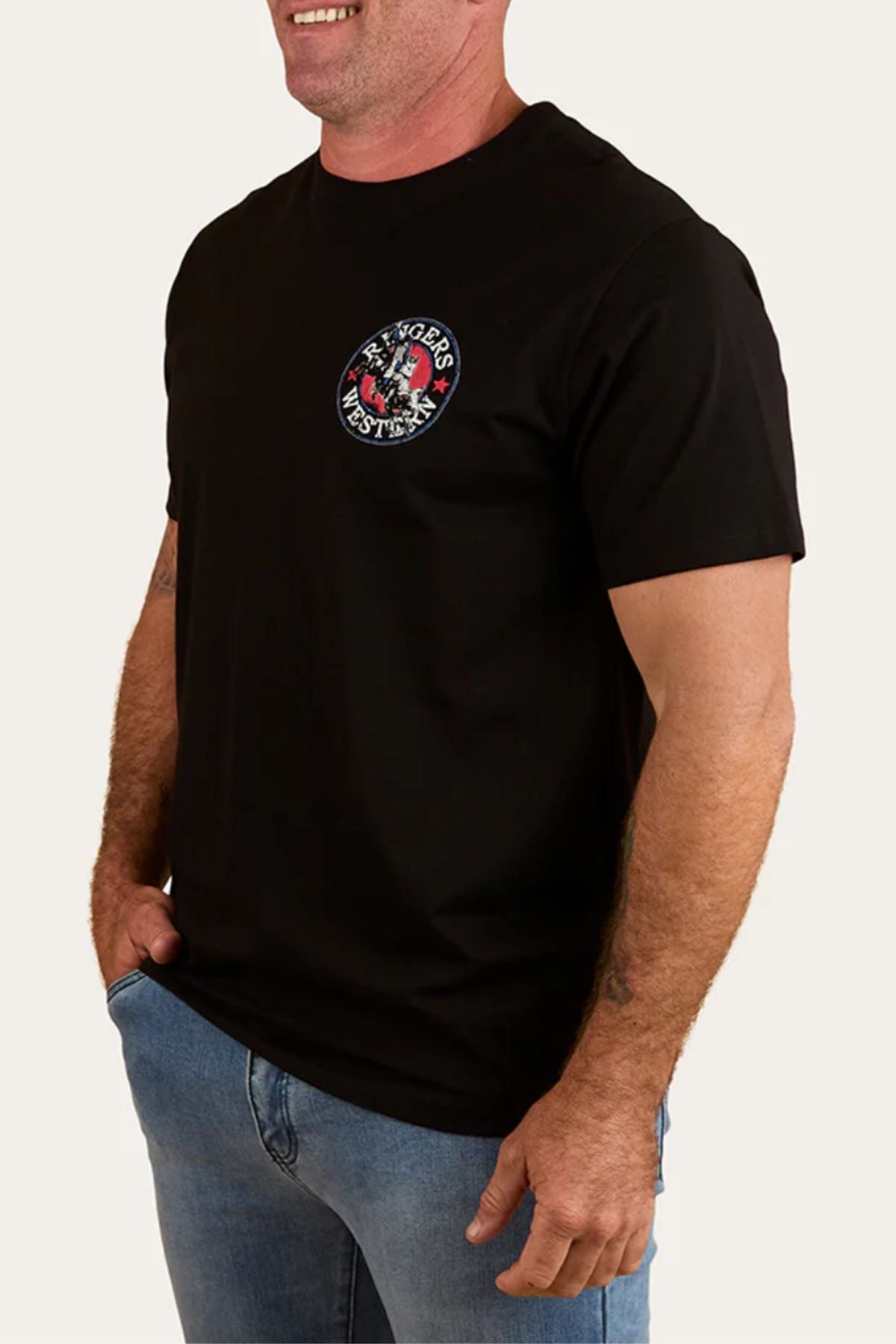 A smiling man wearing the Ringers Western Bucking Bull Loose T-Shirt with a round logo on the chest and blue jeans stands with one hand in his pocket against a plain background.