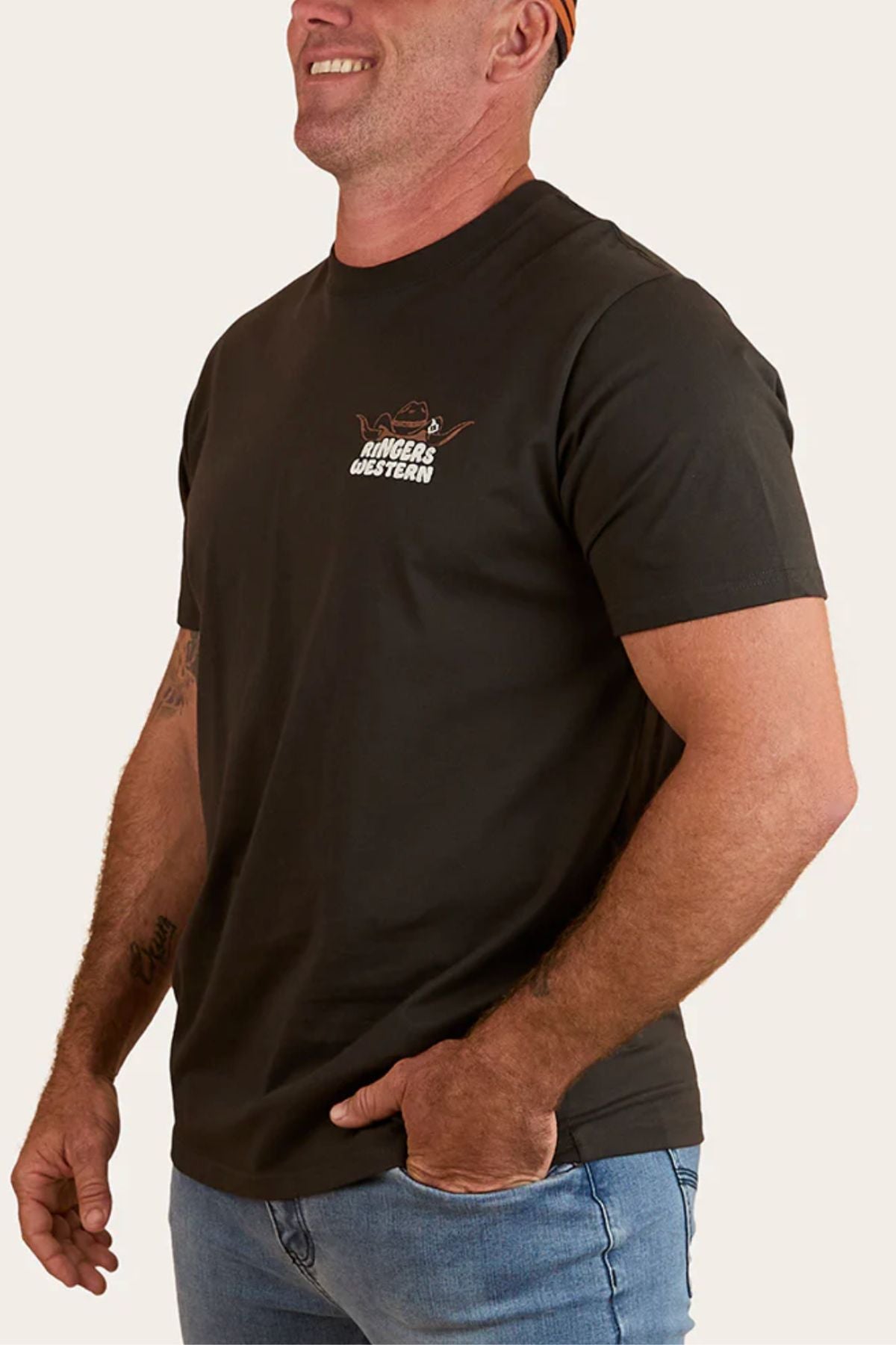 A man smiling and looking to the side wears a black Ringers Western Country Club Loose Fit T-Shirt with a small chest logo and light blue jeans.