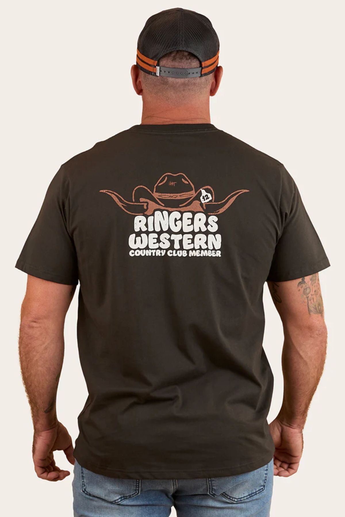 A man, seen from behind, wears a Ringers Western Country Club Loose Fit T-Shirt and cap. The tee has a cowboy hat with horns graphic and "RINGERS WESTERN COUNTRY CLUB MEMBER" text on the back.