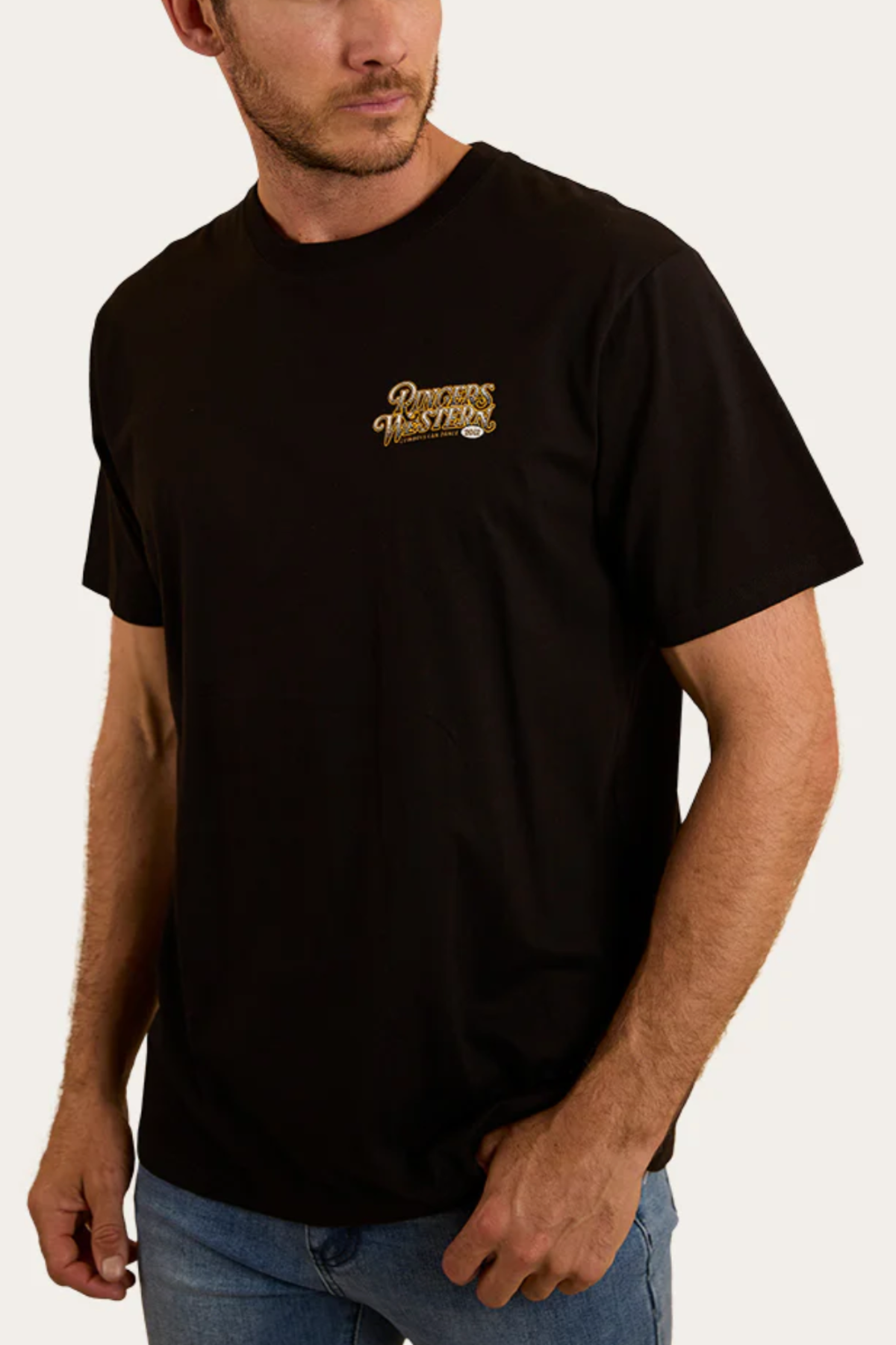 A man stands against a plain, light background wearing blue jeans and a black Ringers Western Cowboys Can Dance Loose Fit T-Shirt with a small gold and white "Records Western Co." logo on the left chest.
