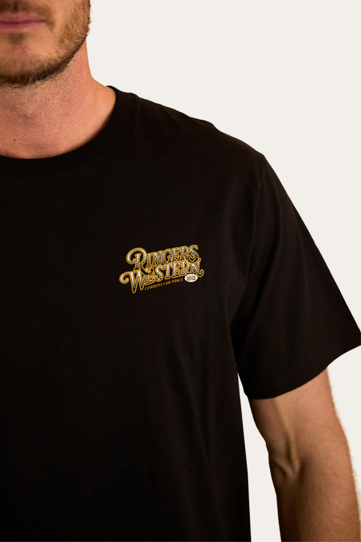A man models the Ringers Western Cowboys Can Dance Loose Fit T-Shirt, featuring a yellow logo on the left chest. Only his lower face and upper torso appear against a plain background, showcasing classic Cowboys Can Dance style.