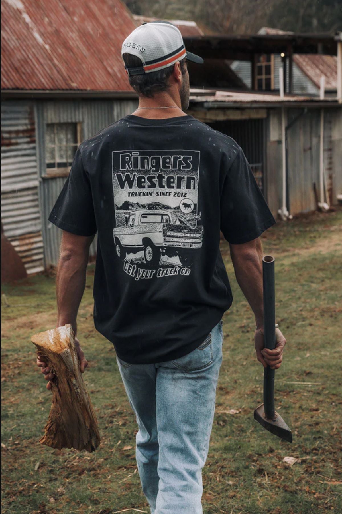 A man in a Ringers Western Get Your Truck On Loose Fit T-Shirt and a cap walks on grass, carrying firewood and an axe, with old metal sheds in the background.