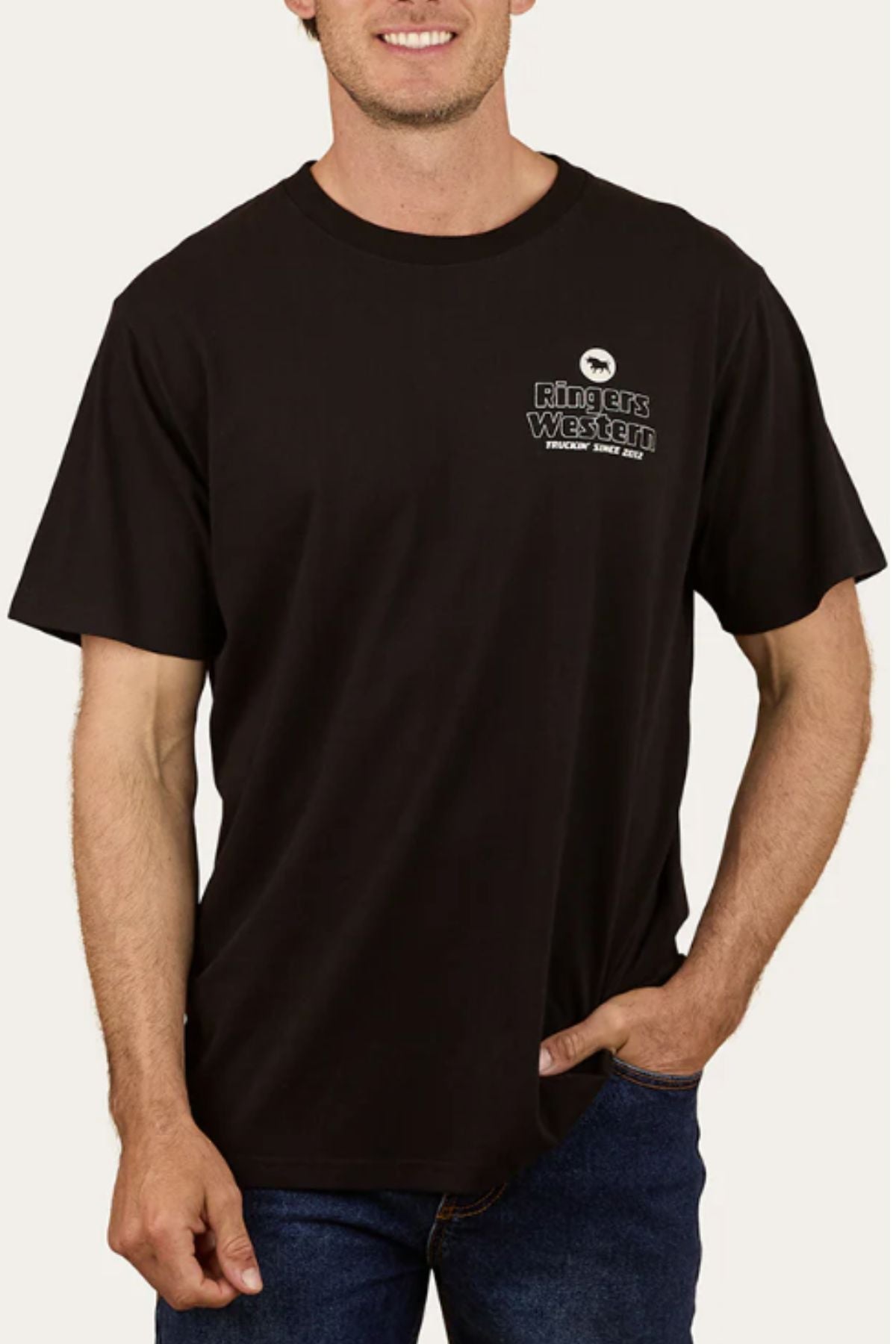 A man wears the Ringers Western Get Your Truck On Loose Fit T-Shirt, made from 100% black cotton with "Ringers Western" and a small left chest logo, paired with blue jeans, standing with one hand in his pocket.