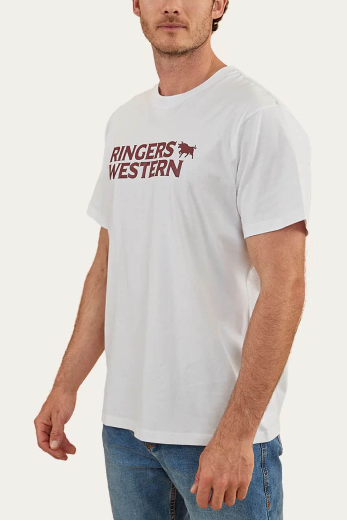 A man wears a Ringers Western Hastings Loose Fit T-Shirt crafted from 100% cotton jersey, marked with "Ringers Western" in maroon and a small horse logo. He pairs it with blue jeans against a plain, light-colored backdrop.