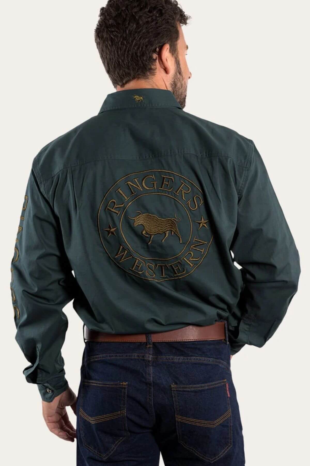 A man is wearing the Ringers Western Hawkeye Embroidered Shirt in dark green premium cotton, featuring "Ringers Western" and a bull logo on the back and left sleeve. His look is completed with dark jeans and a brown belt, viewed from behind.