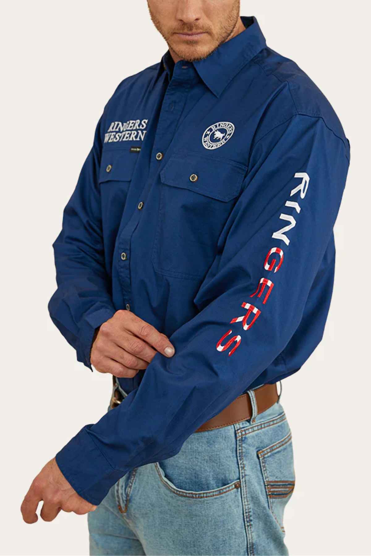 Dressed in a Ringers Western Hawkeye Flag Embroidered Shirt, proudly displaying "RINGERS WESTERN" on the chest and "RINGERS" on the sleeve, a person adjusts their cuff. This shirt, complete with the company logo, pairs perfectly with light blue jeans.