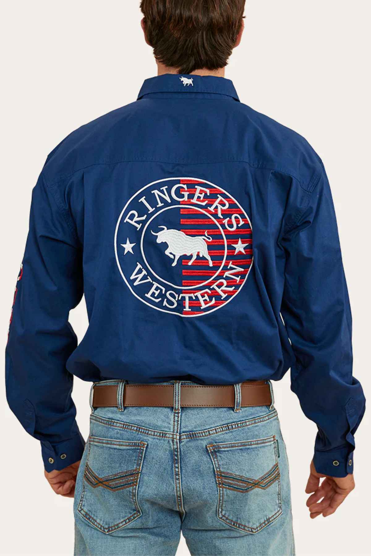 Wearing the Ringers Western Hawkeye Flag Embroidered Shirt, featuring a bull logo and red and white details, the individual complements it with blue jeans and a brown belt. The iconic shirt adds character to this stylish ensemble as they pose with their back to the camera.