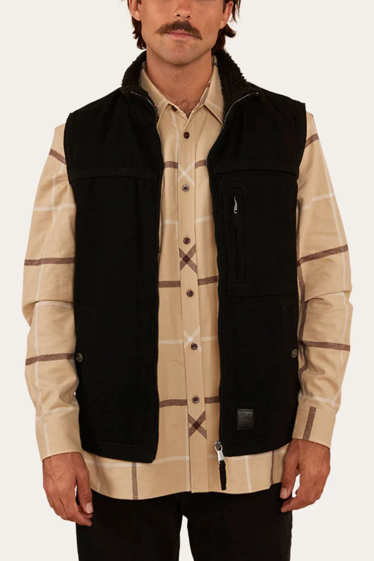 A person with a mustache is wearing a beige plaid button-up shirt under the Ringers Western High Country Vest, which is crafted from durable cotton canvas and features sherpa lining, multiple pockets, and both zipper and button details. The individual has their arms relaxed at their sides against a light gray background, poised for an outdoor adventure.