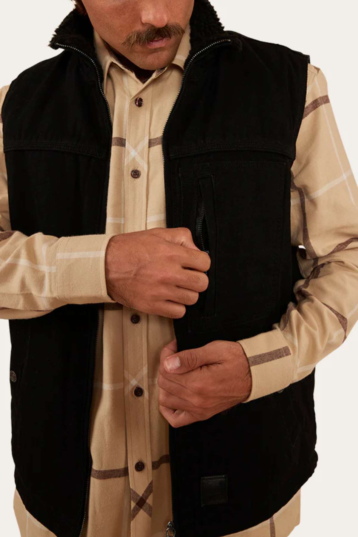 A person wearing a durable cotton canvas plaid shirt and the Ringers Western High Country Vest is shown zipping or unzipping one of the vest's pockets. The person has a mustache and is looking downwards, ready for their next outdoor adventure.