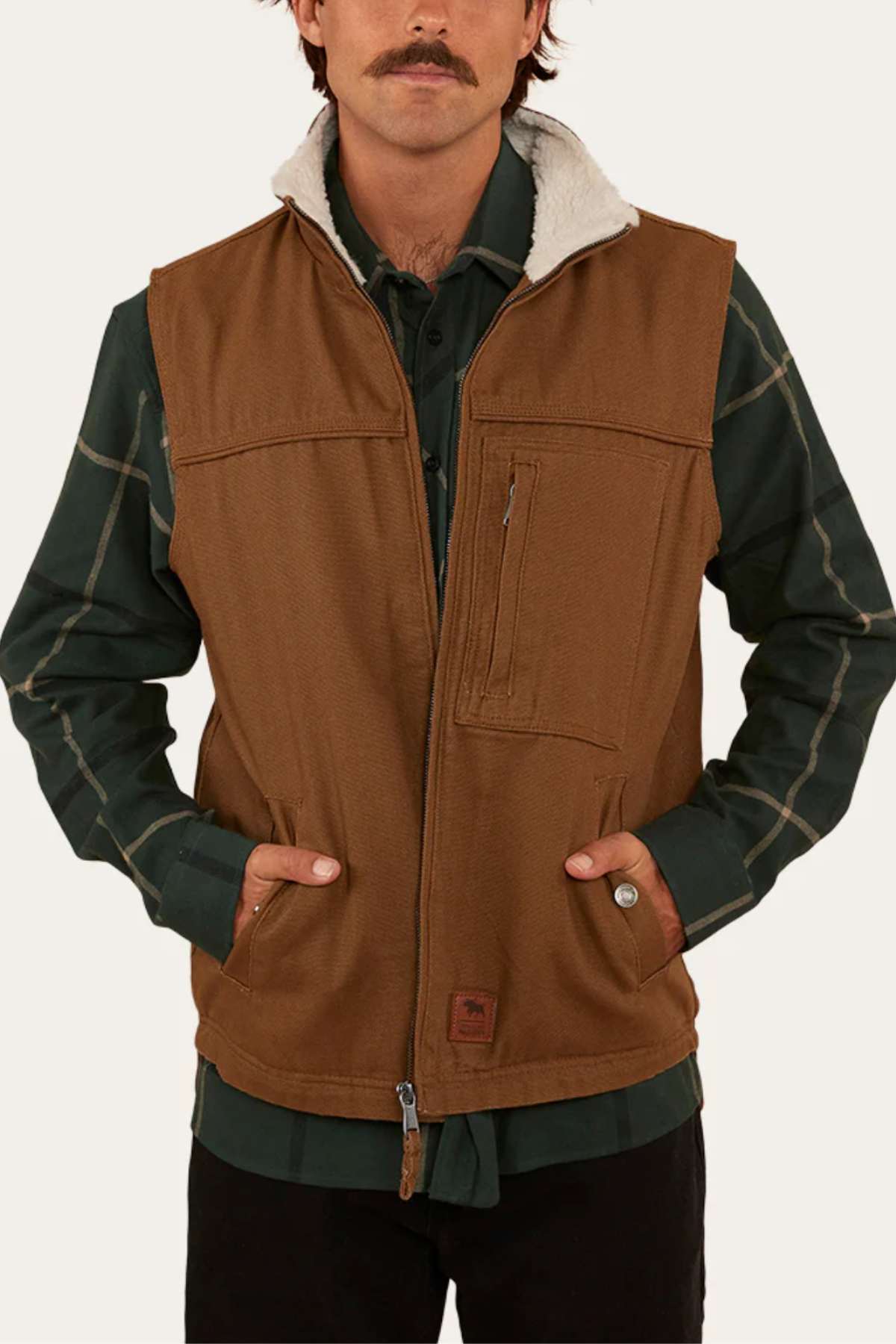 A man with a mustache wears the Ringers Western High Country Vest, featuring cotton canvas fabric and a fleece collar, over a dark green plaid shirt. He stands with hands in vest pockets against a plain off-white background.