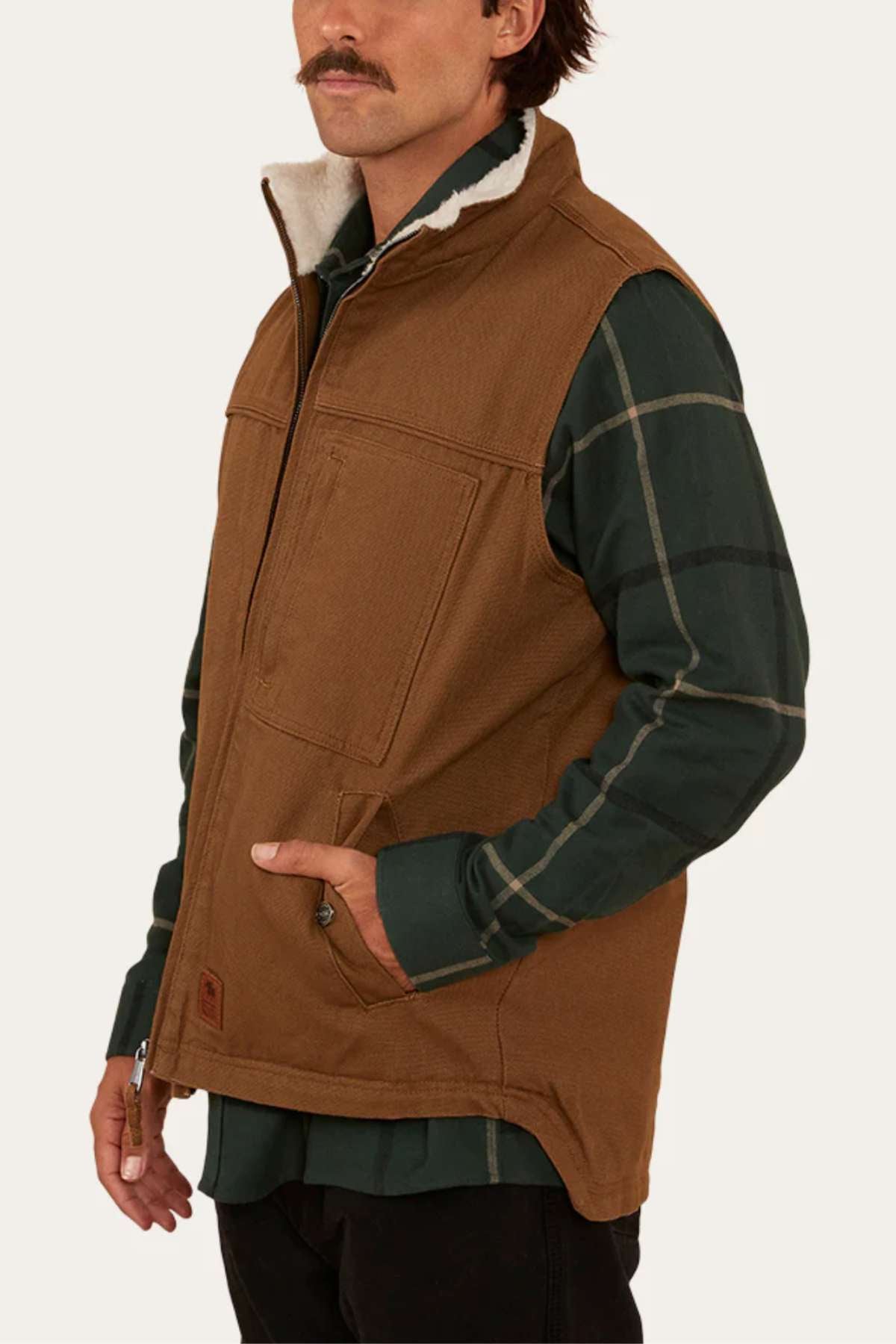A man with a mustache wears the Ringers Western High Country Vest—a brown cotton canvas vest with front pockets and a light-colored lining at the collar—over a green plaid shirt, standing against a plain, light background.