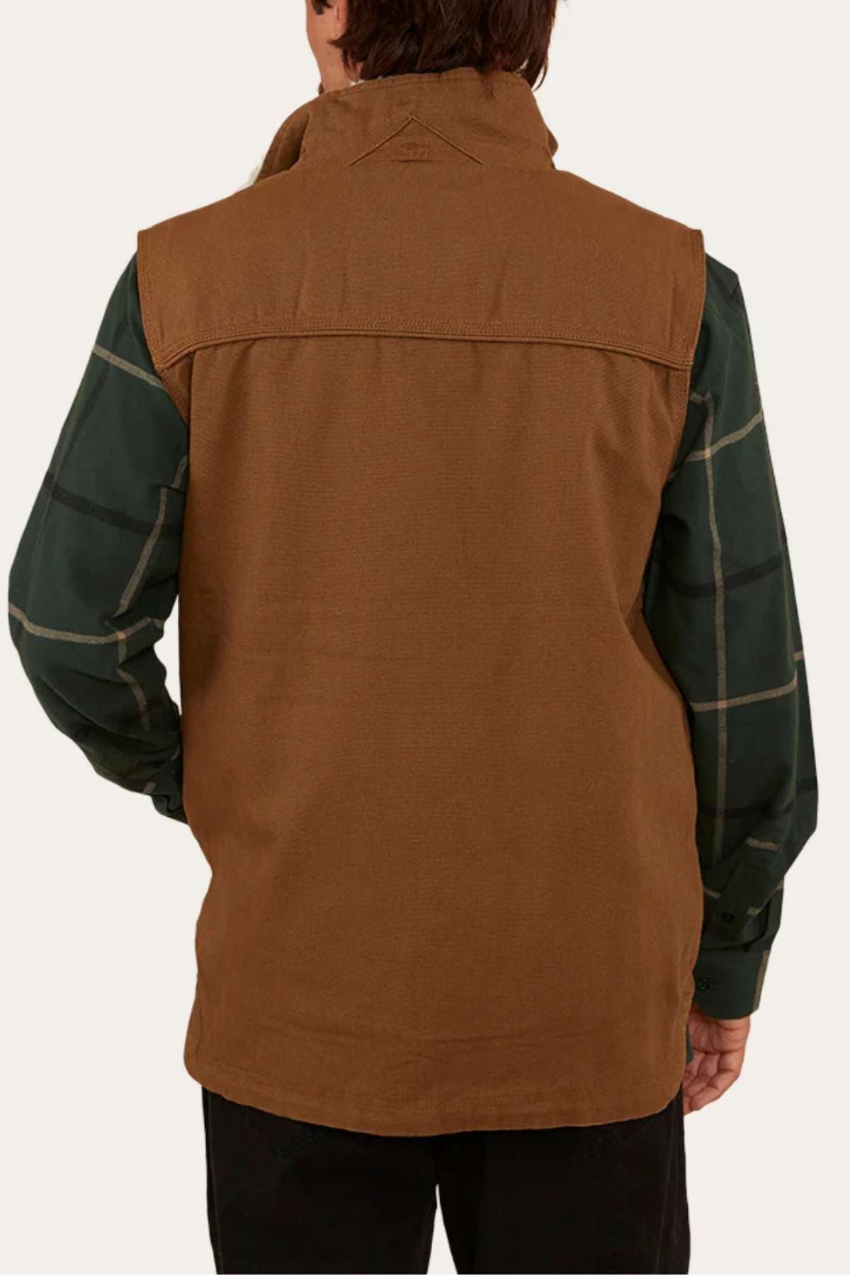A person facing away wears the Ringers Western High Country Vest in brown cotton canvas over a green plaid long-sleeve shirt and black pants, against a plain light-colored background.