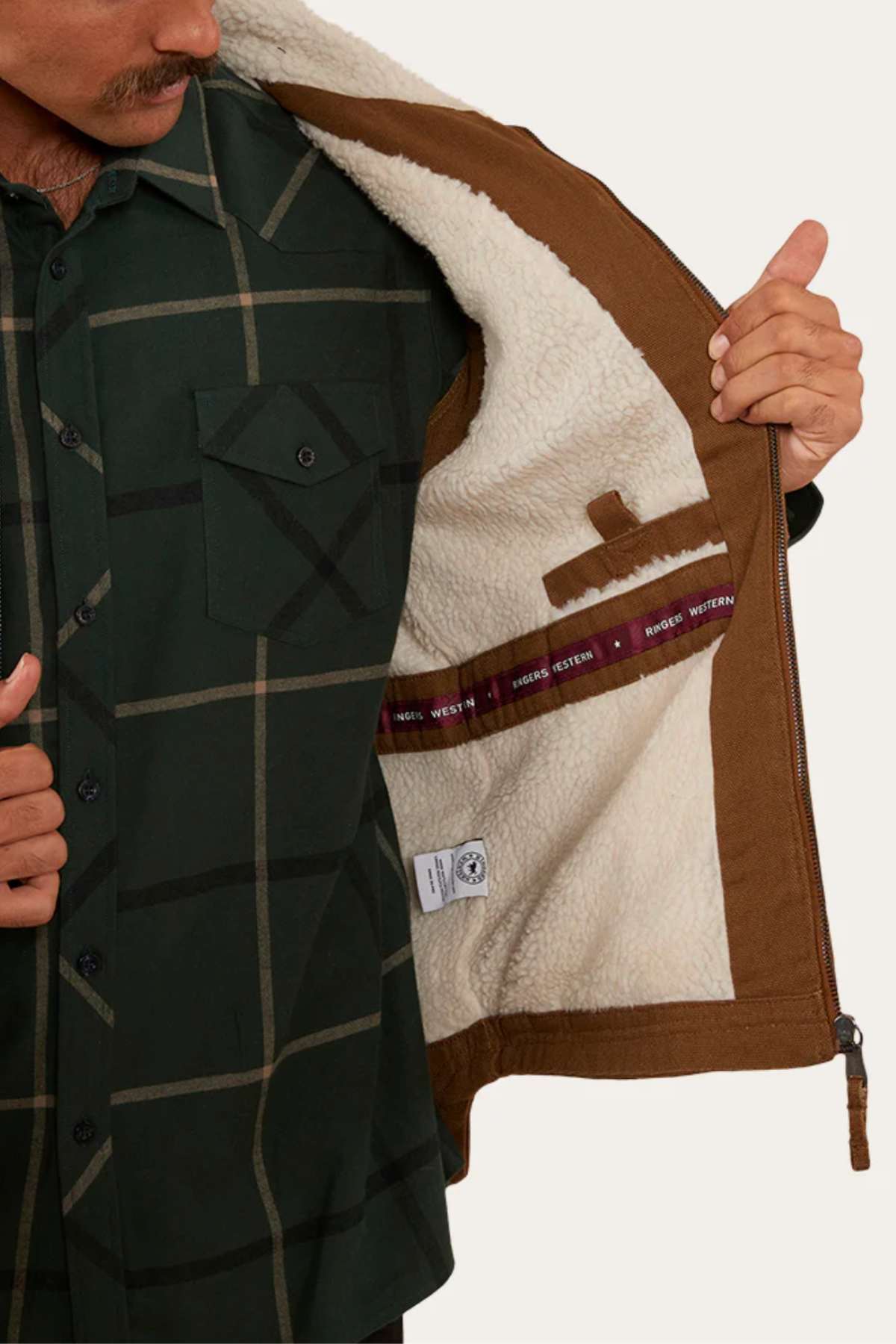 A person wearing a dark green plaid shirt opens the Ringers Western High Country Vest, made of brown cotton canvas with a white fleece lining, showing the inner details and labels.