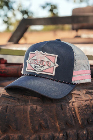 A Ringers Western Hometown Trucker Cap in navy blue and white with a pink patch rests on a dusty tire outdoors, featuring an adjustable snapback and blurred trees and equipment in the background.
