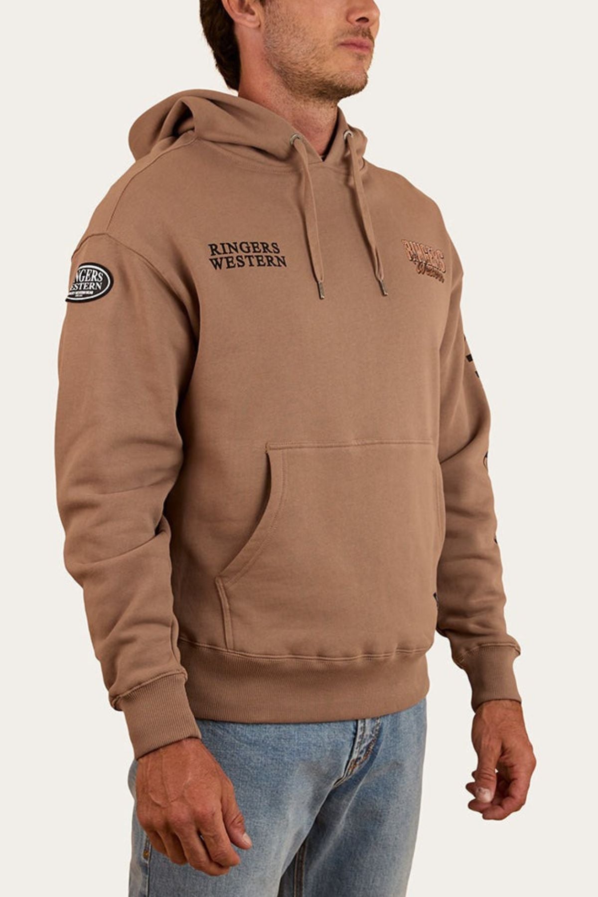 A man stands with arms relaxed, wearing the Ringers Western KNX Oversized Hoodie in brown with chest and sleeve logos, paired with light blue jeans against a plain background.