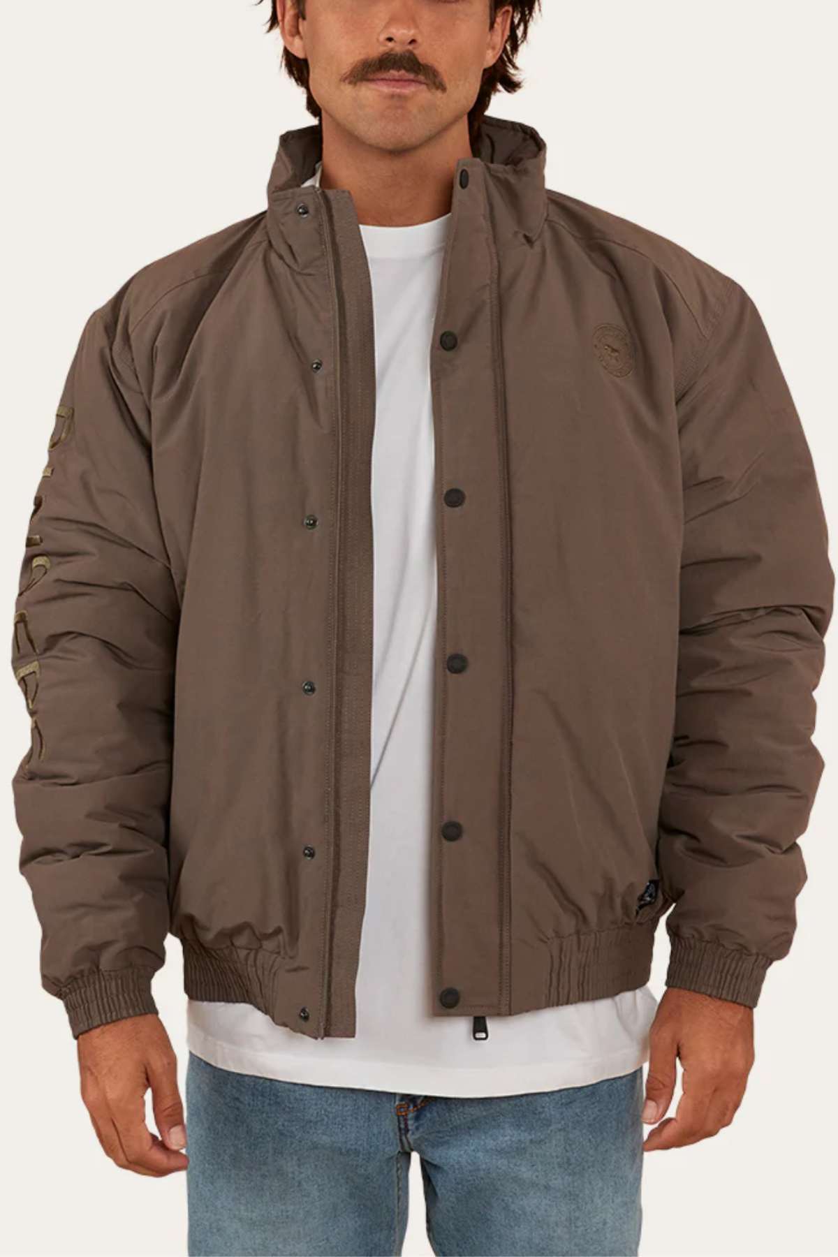 A person stands facing forward wearing a brown, high-collar, button-up Ringers Western Kidman Jacket over a white T-shirt. The jacket, perfect for an outdoor adventure, features elastic cuffs and a subtle emblem on the left chest. The person is also wearing light blue jeans and has short, dark hair and a mustache.