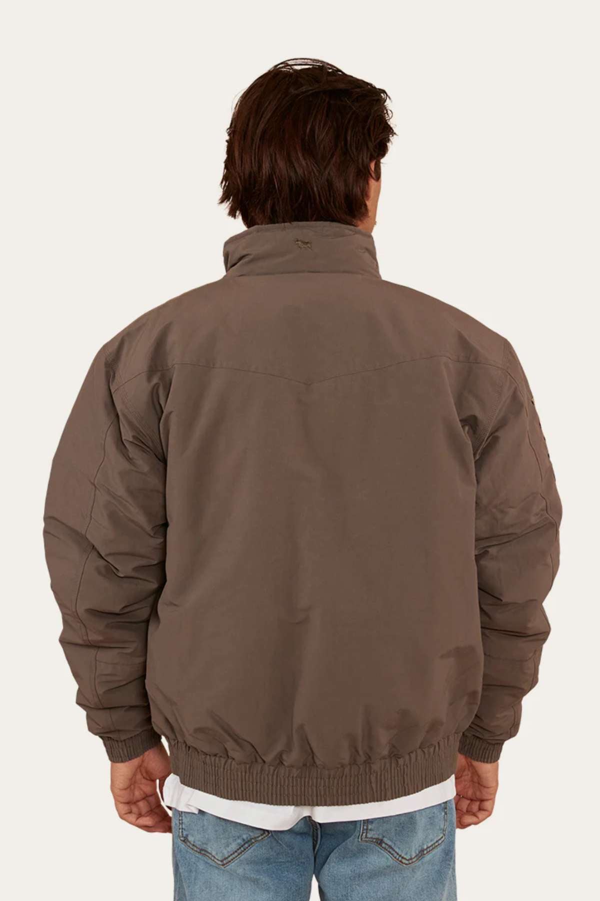 A person with short brown hair is seen from the back wearing a Ringers Western Kidman Jacket, characterized by its brown color, high collar, ribbed cuffs, and quilted lining—ideal for an outdoor adventure. They are also wearing light blue jeans. The background is plain and light-colored.