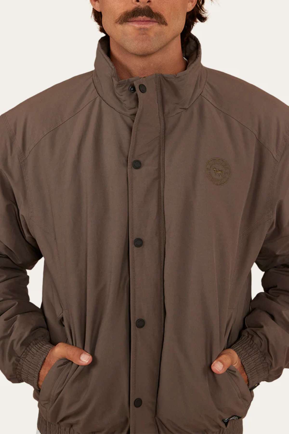 A person with a mustache is wearing a buttoned-up Ringers Western Kidman Jacket in brown, complete with quilted lining, their hands tucked into the pockets. This jacket features ribbed cuffs, a high collar, and a circular logo on the left chest area. Ideal for outdoor adventures, the background is plain white or light-colored.