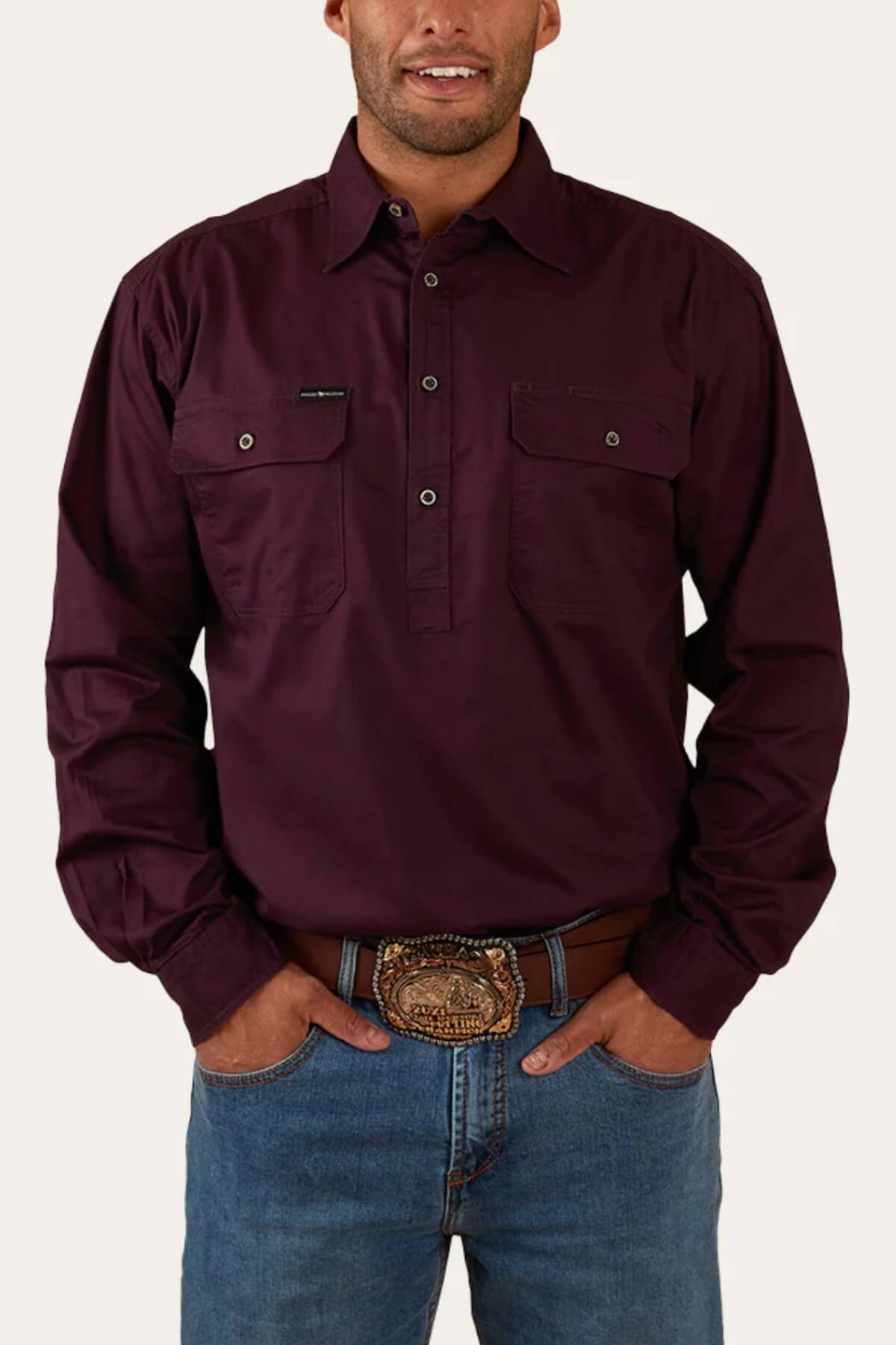 A person wearing a dark maroon Ringers Western King River Half Button Shirt with two chest pockets and denim jeans. They have their hands in their pockets and are also sporting a brown belt with an ornate, oval-shaped buckle. The background is a plain light color.