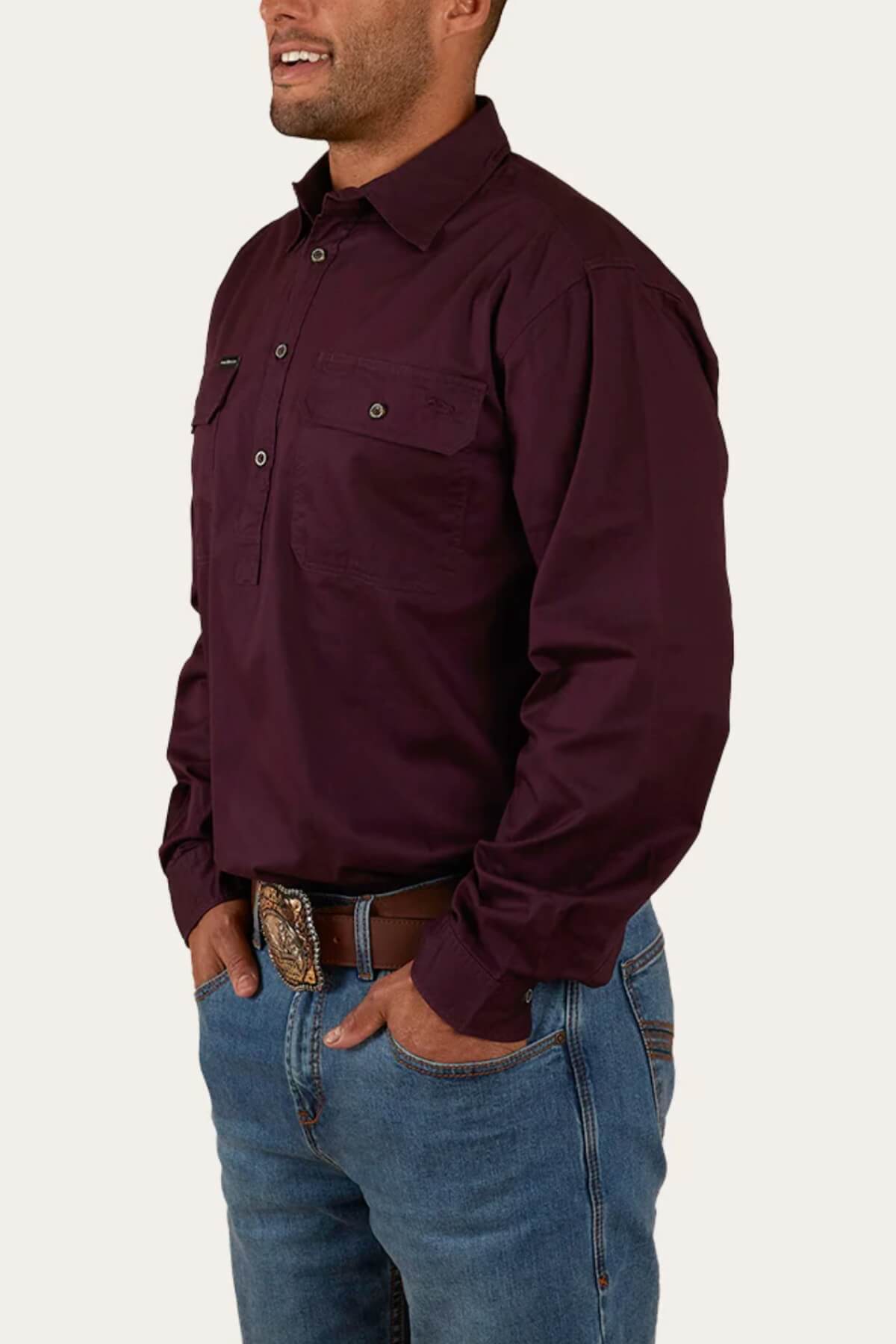 A man stands with his hands in the pockets of his blue jeans, wearing a long-sleeved dark maroon Ringers Western King River Half Button Shirt. He accessorizes with a decorative belt featuring a prominent buckle. The plain white background keeps the focus on his attire.