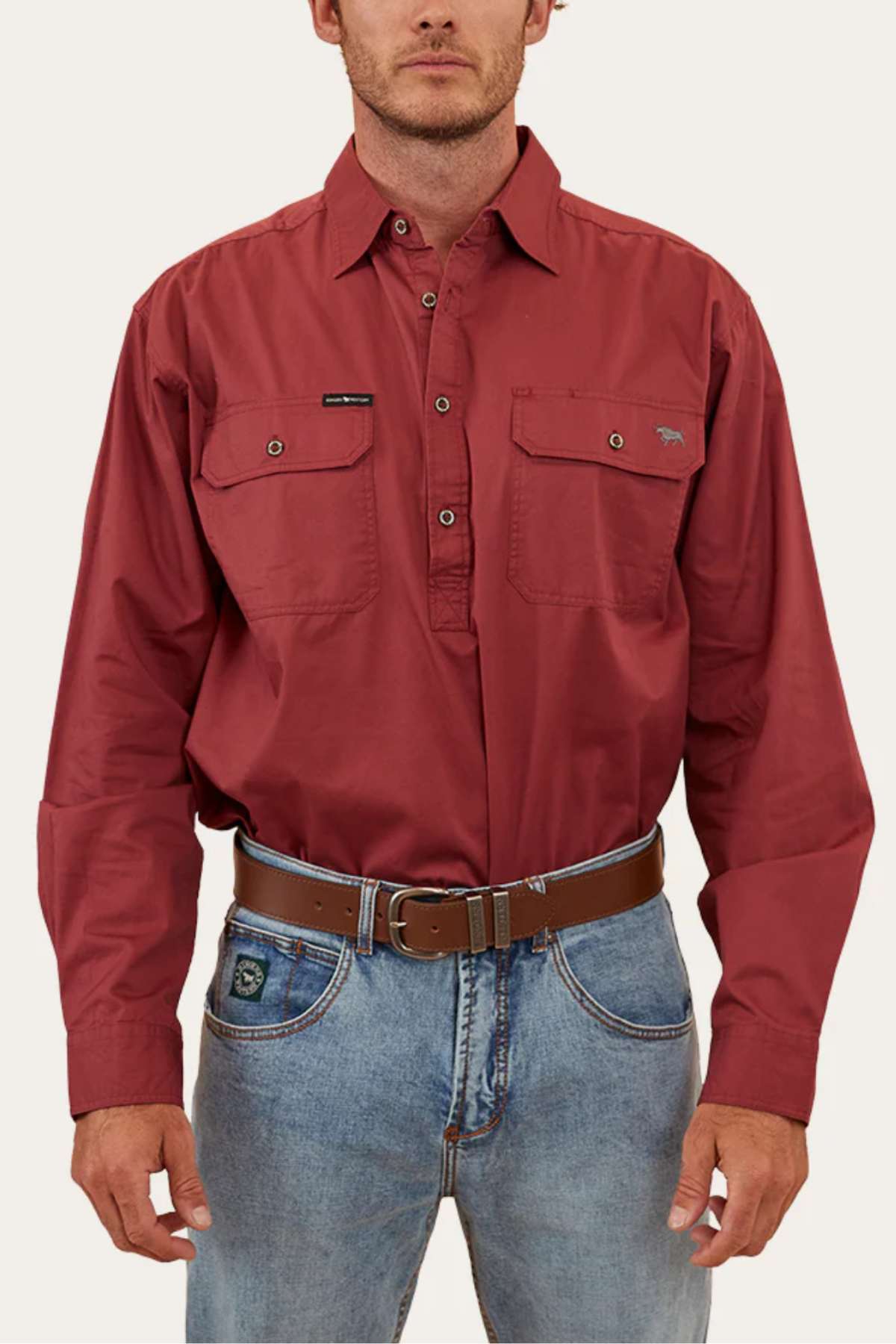 A man stands against a plain background wearing a Ringers Western King River Half Button Shirt, long-sleeved and maroon, paired with faded blue jeans. The shirt features two chest pockets and is made of 100% cotton for durability and comfort. He accessorizes with a brown belt featuring a silver buckle, with his hands resting by his sides.