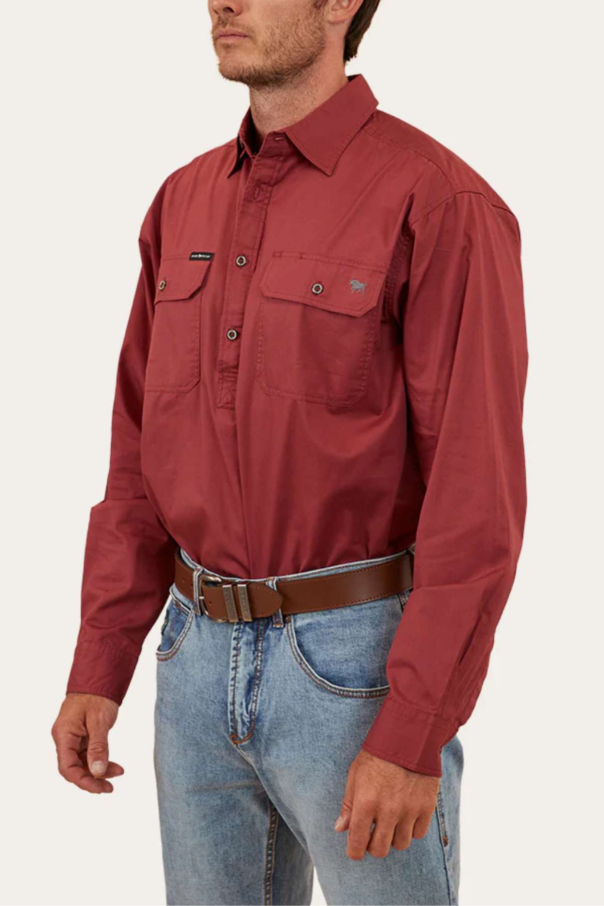 A person wearing a maroon Ringers Western King River Half Button Shirt with two front pockets, light blue jeans, and a brown belt stands against a plain backdrop. The person’s face is not visible.