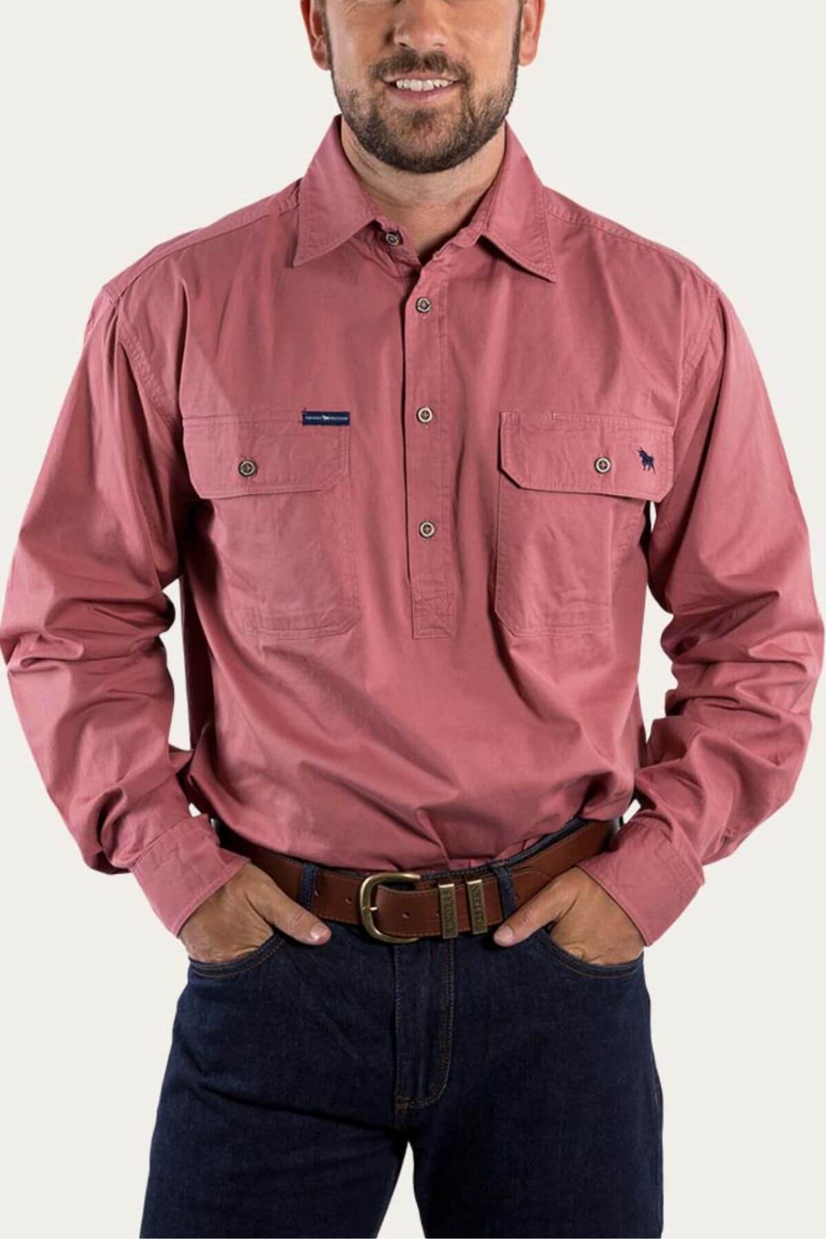 A person stands with hands on hips, wearing a Ringers Western King River Half Button Shirt, which is a comfortable, long-sleeved button-up pink work shirt made from 100% cotton featuring two chest pockets. The shirt is paired with dark jeans and a brown belt against a plain, light-colored background.