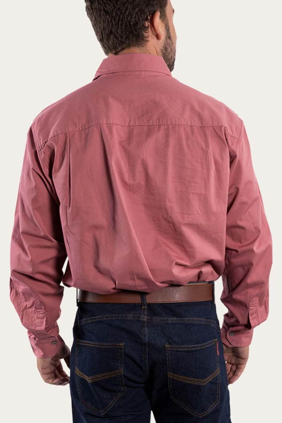 A man with short dark hair stands facing away, wearing a long-sleeved Ringers Western King River Half Button Shirt in red, tucked into dark blue jeans. The 100% cotton shirt features buttoned cuffs, and he is accessorized with a dark brown belt. The background is plain and light-colored.