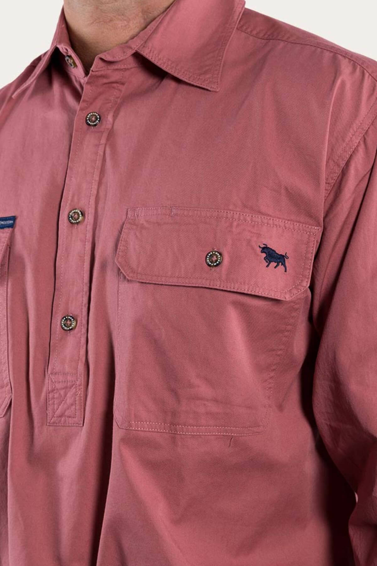 Close-up of a person wearing the Ringers Western King River Half Button Shirt in a muted reddish-pink color. This comfortable, 100% cotton long-sleeve work shirt features a chest pocket with a buttoned flap and an embroidered small blue animal silhouette, likely a horse, above the pocket.