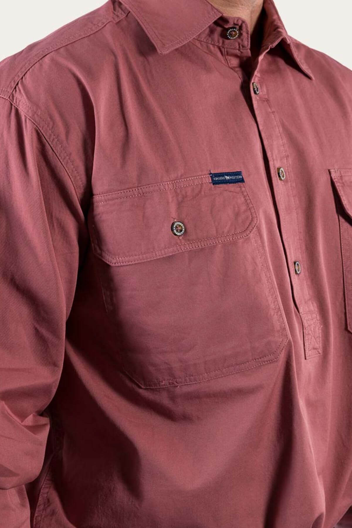 A person wearing a Ringers Western King River Half Button Shirt in a muted red color. Made from 100% cotton, the comfortable long-sleeve shirt features two chest pockets, one adorned with a small label containing text. Only the torso and part of the upper arm are visible.
